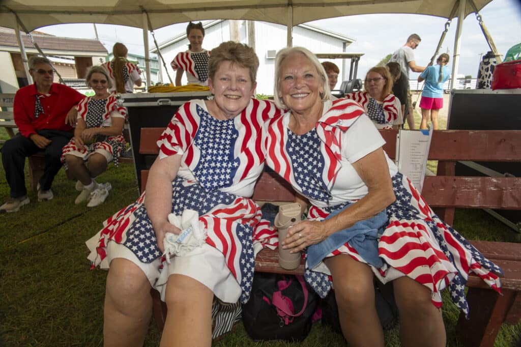 Two women in patriotic red, white, and blue outfits sit smiling under a tent, enjoying a fair or community event.