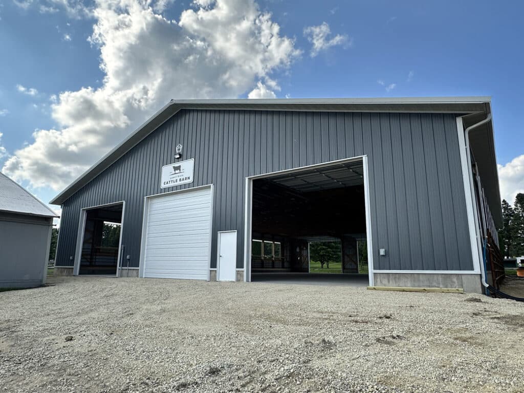 A newly built goat barn with large overhead doors stands on the fairgrounds, featuring a clean, modern design under a cloudy sky.