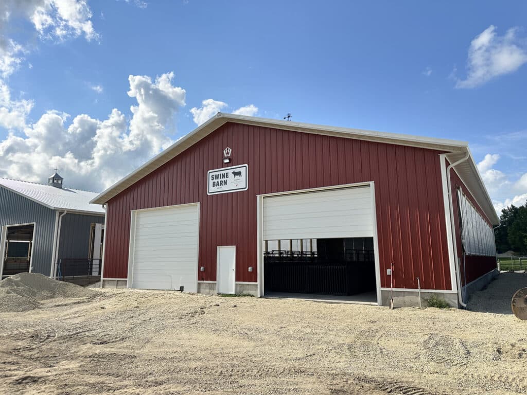 A newly constructed swine barn with a red exterior and large overhead doors sits on the fairgrounds under a bright, sunny sky.