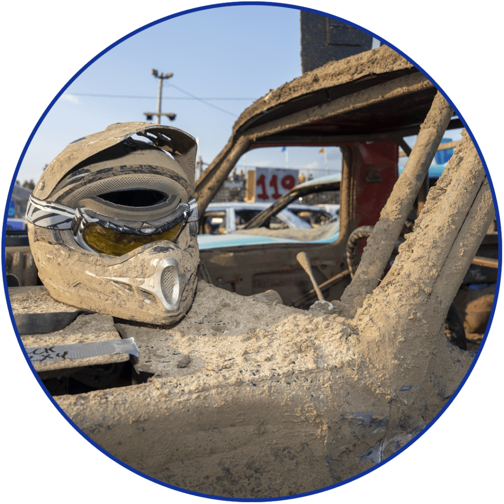 Muddy racing helmet resting on a dirt-covered demolition derby car at an outdoor event.